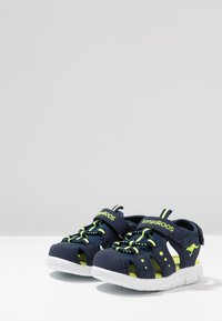 Navy blue sandals with green accents, mesh upper, adjustable strap, and white outsole. Features cutouts for ventilation and flexibility.