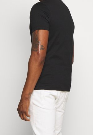 Homme portant une chemise noire à manches courtes et un pantalon blanc, montrant un tatouage de papillon et une cicatrice sur son bras supérieur gauche.