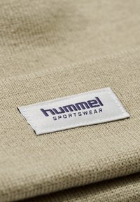 Beige stof met een gestructureerd oppervlak met een wit label. Het label toont het merk "hummel" in paarse tekst, dat sportkleding aangeeft.