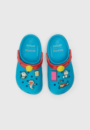 Sinised plastikust sussid punaste rihmadega, millel on multikangelaste kleebised ja dekoratiivsed aksessuaarid nagu kell ja uks, ning millel on perforatsioonimuster.