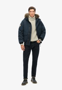 Mørkeblå pufferjakke med pelsforet hætte, grå turtleneck sweater, mørke jeans og strukturerede grå støvler med rund tå.