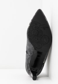 Chaussure à talon haut texturée noire pointue, inclinée pour montrer la semelle avec des motifs en relief de pièces de monnaie et la marque "Peter Kaiser".