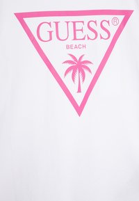 Bílý bavlněný tričko s velkým růžovým trojúhelníkovým logem a textem "GUESS BEACH" uprostřed s designem palmy.