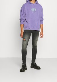 Sudadera morada con bolsillo frontal y diseño gráfico, combinada con jeans negros desgastados y botas negras hasta el tobillo con suela gruesa.