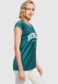 Teal jersey t-shirt med korte ærmer og v-hals, med hvid tekst "BERLIN" og "MERCH CO." nedenunder. Kombineret med hvide bukser.