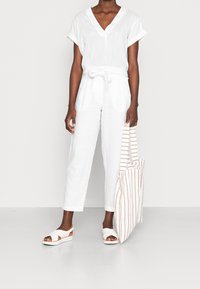 Top in lino bianco con scollo a V, abbinato a pantaloni in lino bianco. La modella tiene in mano una borsa a tracolla a righe bianche e beige e indossa sandali bianchi.