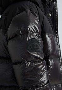 Giacca a vento nera con finitura lucida, texture trapuntata e una toppa in gomma con logo sulla manica. Taschini con zip visibili.