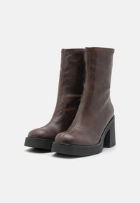 Braune Stiefeletten aus Kunstleder mit glatter Textur, quadratischer Zehenform, hohem Blockabsatz und kontrastierenden Nähten.