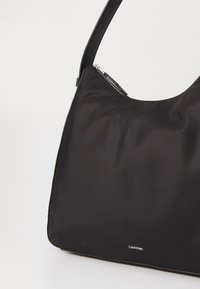 Czarna nylonowa torebka o gładkiej fakturze, zaokrąglonym kształcie i pojedynczym pasku na ramię. Posiada zamknięcie na suwak oraz małe logo.
