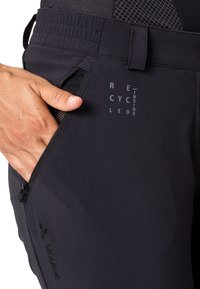 Crne biciklističke hlače s patentnim zatvaračem na džepu, teksturirana tkanina i otisak "RECYCLED INSIDE". Imaju elastični pojas i suptilan brend.