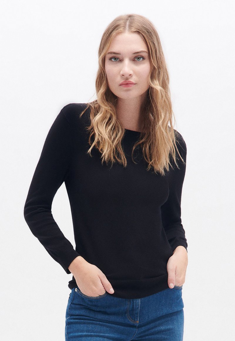 Zwarte lange mouw sweater in een aansluitend ontwerp, met een gladde textuur en ronde hals. Gecombineerd met blauwe jeans staat het model voor een neutrale achtergrond.