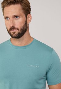 Camiseta de manga corta color turquesa hecha de una tela suave. Presenta un cuello redondo y un pequeño logo blanco "FACONNABLE" en el pecho.