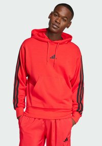 Felpa rossa con strisce nere sulle maniche, tasca anteriore e cappuccio regolabile con cordino. Realizzata in materiale morbido, presenta il logo Adidas sul petto.
