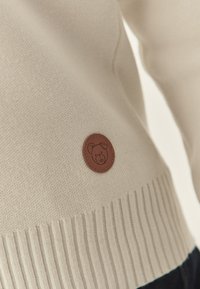 Pulover tricotat de culoare crem, cu tiv striat și opată rotundă din piele maro, care prezintă un design cu un urs pe partea stângă jos.