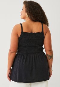 Zwarte tanktop met een gesmokte lijf, dunne bandjes en een uitlopende zoom. De stof lijkt lichtgewicht met een subtiele glans. Heeft een vlinder tatoeage.