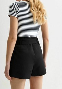 Shorts noirs taille haute au design ajusté, avec une texture lisse et deux poches arrière. Haut côtelé rayé partiellement visible.