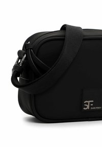 Czarna torebka na ramię wykonana z gładkiego materiału, o usztywnionym kształcie, szerokim pasku z tkaniny oraz metalicznym logo po boku.