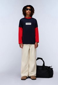 Tamsiai mėlynas trumpomis rankovėmis marškinėlis su raudonos spalvos ilgomis rankovėmis marškinėliais po apačia, smėlio spalvos plačios kelnės, rudi batai ir juoda sportinio stiliaus kuprinė. Juoda kibiro formos kepurė ir akiniai nuo saulės.