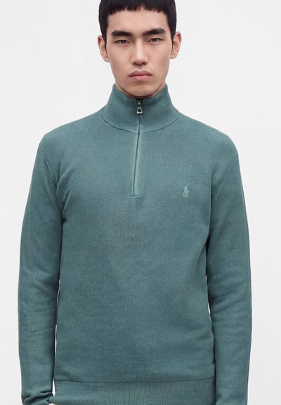 Jeune homme portant un pull en maille bleu sarcelle avec une fermeture éclair quart de zip et un col montant, debout devant un fond clair uni.