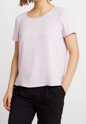 Kvinde iført en lyserød kortærmet bluse med rund halsudskæring og sorte bukser, stående med den ene hånd i lommen.