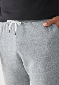 Grijze joggingsbroek van zacht materiaal, met een verstelbare tailleband met witte koorden en metalen uiteinden; zijzakken zijn zichtbaar.
