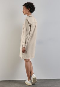 Robe chemise en lin beige avec col, manches longues et poignets boutonnés, portée avec des baskets blanches sur un fond gris.