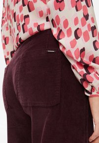 Pantalon en velours côtelé bordeaux avec poches arrière, orné d'une petite étiquette argentée. Associé à un chemisier à motifs dans des tons roses et rouges.