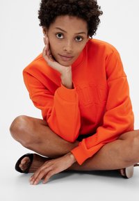 Oranžová mikina s vystouplým logem, volný střih, žebrované manžety, nošena s černými nazouvacími sandály. Osoba sedí v uvolněné pozici.