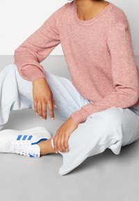 Rosa Strickpullover mit gerippten Bündchen, hellblaue lässig geschnittene Jeans und weiße Sneakers mit blauen Streifen und Schnürsenkeln.
