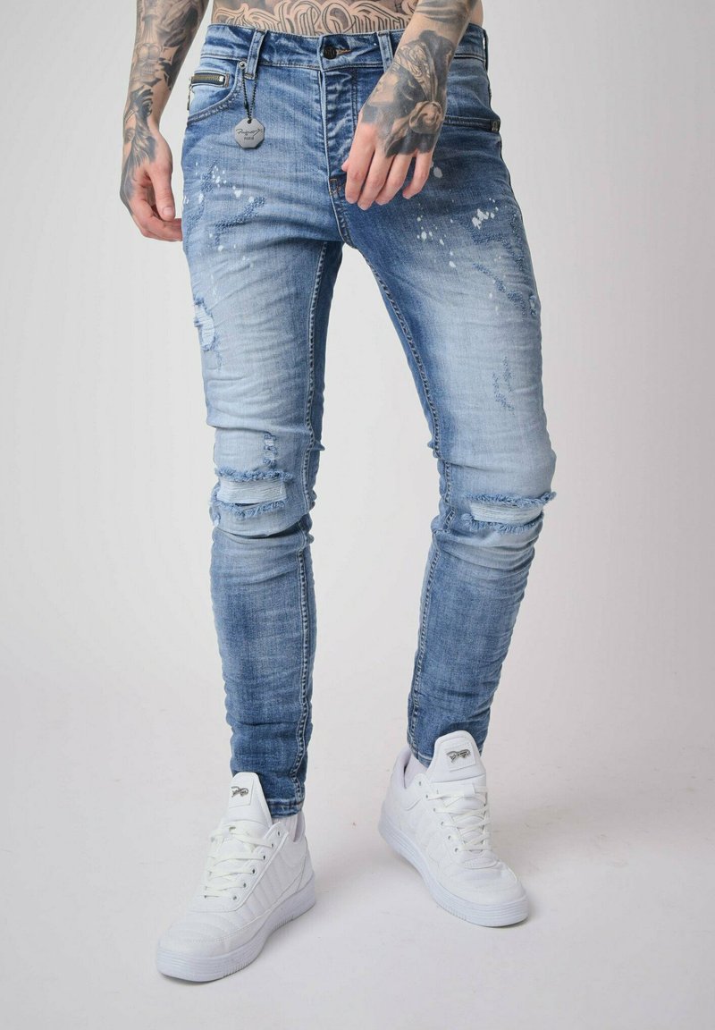 Light blue distressed jeans with rips, tapered fit, and faded texture. Paired with white sneakers, showcasing a casual, contemporary style.