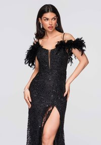 Robe noire en tissu transparent avec un travail de perles et des ornements complexes, accents en plumes sur les épaules, et une fente haute jusqu'à la cuisse.