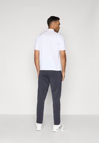 Cămașă polo albă cu mâneci scurte și spate texturat, asortată cu pantaloni sport gri închis cu un croi slim și un buzunar cu fermoar, purtată cu adidași albi.