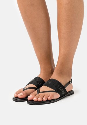 Pieds portant des sandales plates noires avec de larges brides en tissu et de fines brides à l'orteil, avec le texte "CALVIN JEANS" imprimé sur la bande en tissu.