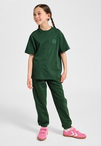 Grøn bomulds T-shirt med et lille lomme-logo, parret med matchende grønne joggers. Modellen har lyserøde sneakers med velcro-stropper på.