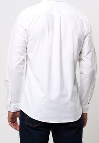 Biała koszula z długim rękawem z szwem na tylnej części, mankietami na guziki i zaokrąglonym dołem, zestawiona z ciemnymi jeansami.