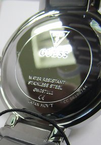 Baksidan av en klocka i rostfritt stål med ingraverad logotyp "Guess", modelluppgifter, vattentålighet och information om rörelse. Svart etikett fäst.