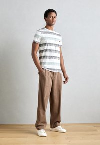 Camiseta de rayas de mangas cortas en blanco, gris y verde, combinada con pantalones marrón claro. Zapatillas blancas completan el look.