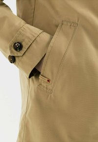 Krémová nepromokavá bunda s hladkou texturou, knoflíkovými manžetami a boční kapsou s malou značkovou cedulkou. Viditelné švy.