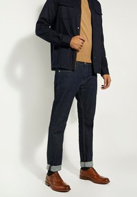 Marineblauwe overhemels, beige trui, donkere spijkerbroek, brown dresschoenen met brogue-details. Opgerolde mouwen onthullen een contrasterende textuur.