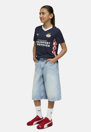 Maglia sportiva della marina con colletto bianco e dettagli rossi, abbinata a pantaloni corti in denim azzurro chiaro e sneakers rosse con calzini bianchi.