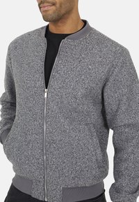 Giacca bomber grigia con tessuto testurizzato, chiusura con zip e polsini a costine. Presenta un colletto grigio uniforme e una vestibilità rilassata, ideale per essere indossata a strati.