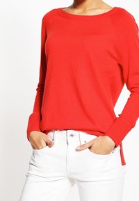 Kvinna klädd i en ljusröd långärmad tröja med vid halsringning och vita högmidjade byxor, med händerna i fickorna.