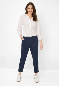 Witte geborduurde blouse met driekwart mouwen, gecombineerd met marineblauwe tapered broek. Model draagt witte sneakers. Eenvoudig ontwerp, neutrale kleuren.