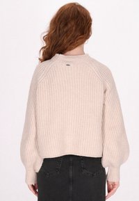 Helles beiges Strickpullover mit strukturierter Musterung, Raglanärmeln und gerippten Bündchen. Enthält ein kleines Logo in der Nähe des Kragens.