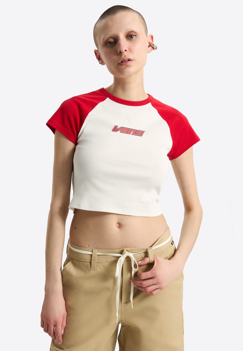 Person mit rasiertem Kopf trägt ein weißes und rotes, verkürztes Vans-T-Shirt und beige Hose mit weißem Kordelgürtel vor einfarbigem Hintergrund.