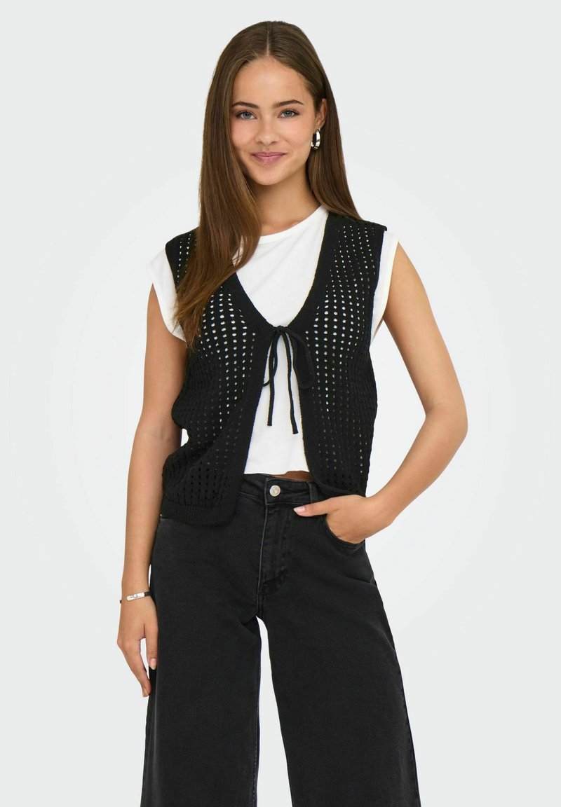Gilet nero a maglia con design a rete, chiusura con laccetto, indossato sopra una camicia bianca e abbinato a jeans neri a vita alta. La modella posa in modo informale.