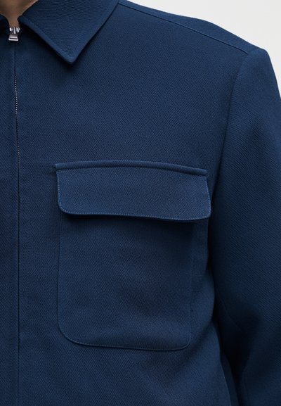 Σκούρο μπλε μπουφάν με υφή, με εμπρόσθιο φερμουάρ και μεγάλη τσέπη στο στήθος με καπάκι στην αριστερή πλευρά.