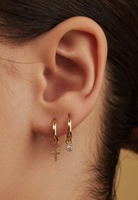 Gold hoop earrings with a small cross charm and a crystal accent, hanging from the lobe, against a neutral background.