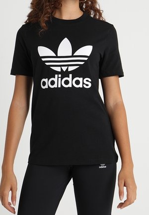 Femme portant un t-shirt noir Adidas avec un logo trèfle blanc et un legging noir Adidas avec un petit logo sur la cuisse gauche.