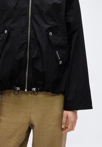 Μαύρο μπουφάν Barbour με φερμουάρ εμπρός, τσέπες με καπάκι και κουμπιά snap, κορδόνι στο τελείωμα, φορεμένο με ανοιχτό καφέ παντελόνι, εμφανές το δεξί χέρι του μοντέλου.